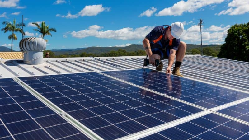 Solar installer wearing a hard hat and safety harness working on the mounting of large solar panels on a low-sloped, metal corrugated roof.